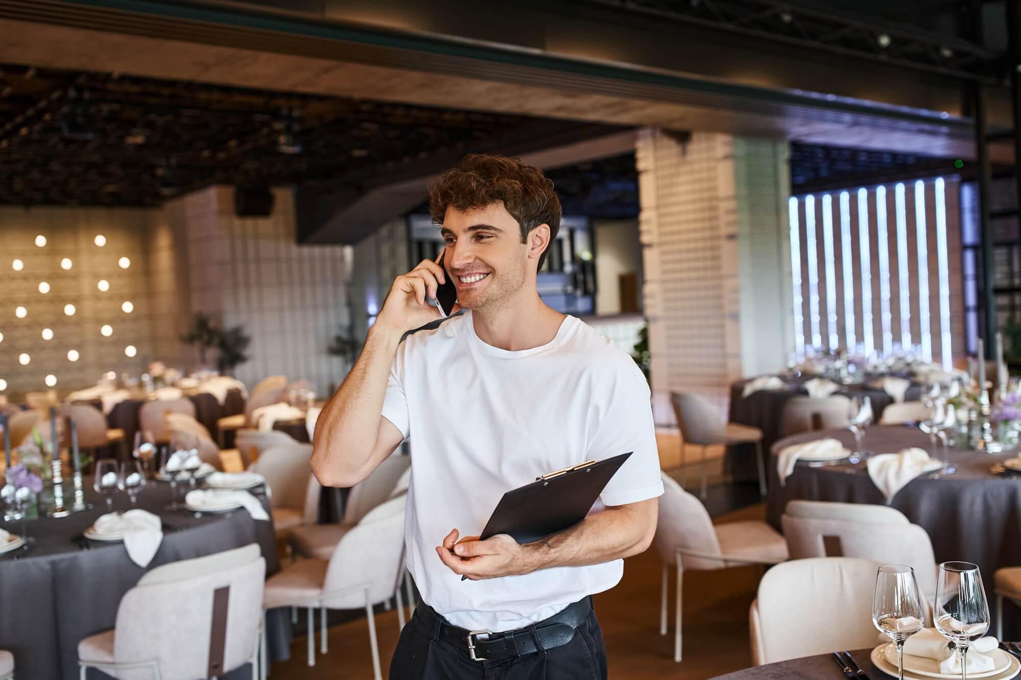 smiling event organizer with clipboard talking on smartphone near festive tables in banquet hall